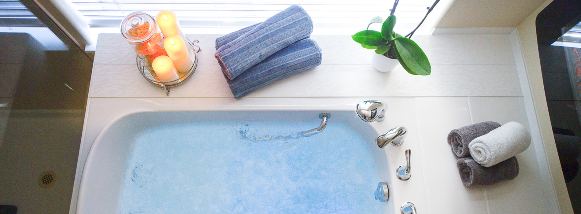 The image shows a modern bathroom interior with a bathtub, a window, a potted plant, and various bathing accessories on the windowsill.