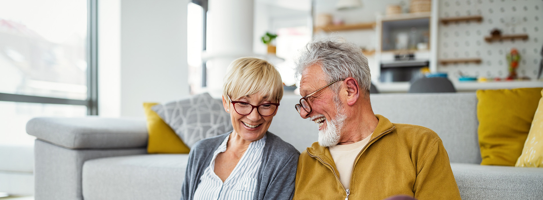 The image depicts two older adults, a man and a woman, sitting together on a couch with a modern interior design.