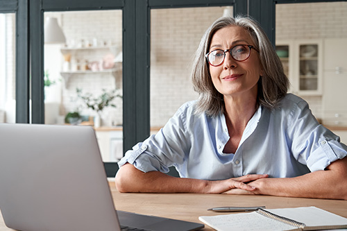 This is a photograph of an older woman seated at a table with her hands clasped together, looking directly at the camera with a neutral expression. She appears to be indoors, possibly in a home setting, with a laptop and some papers on the table.