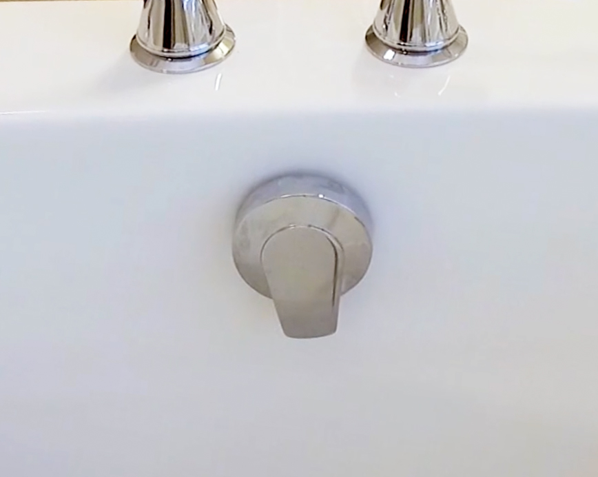 The image shows a close-up of a white porcelain bathroom sink with a silver faucet and handle, positioned against a wall with two visible knobs, one of which appears to be the faucet control.
