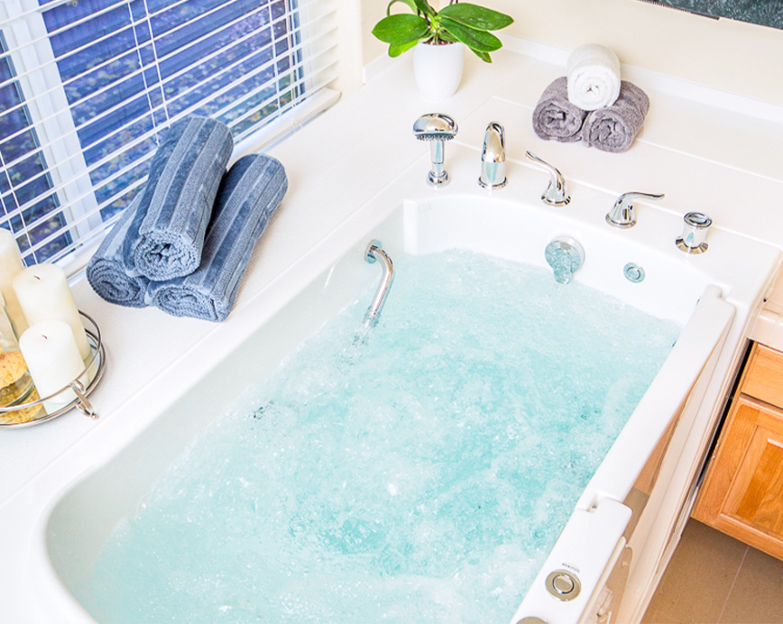 The image shows a modern bathroom interior with a large freestanding bathtub filled with blue water, flanked by rolled-up towels on either side, and a neatly arranged sink area with a mirror above it.