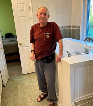 The image shows an older man standing in front of a bathroom sink with his hands on his hips, wearing a black t-shirt and blue pants. He appears to be in a residential setting, possibly a kitchen area due to the presence of a countertop and cabinets.