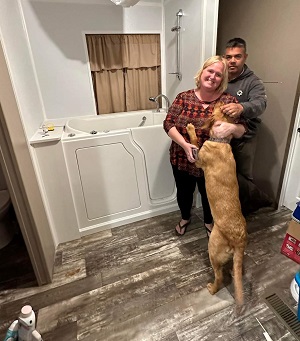 The image depicts a man and woman standing in a bathroom with a dog on a leash, smiling at each other.