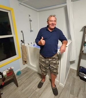 The image shows a man standing in a bathroom, giving a thumbs-up gesture with his right hand while wearing a camouflage patterned hat and a blue shirt.