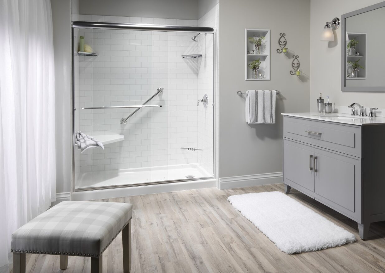 The image shows a modern bathroom with a walk-in shower, a sink vanity, and a bench at the foot of the tub.