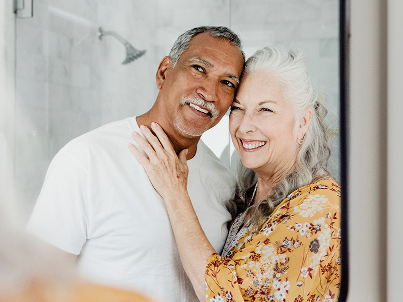 A man and woman are embracing each other with smiles, standing close together in front of a mirror.