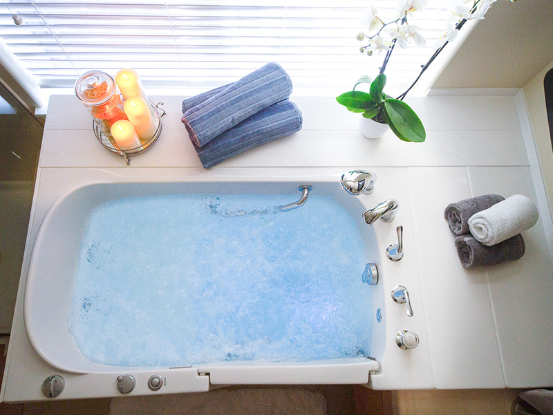 The image shows a bathtub with blue water and a white frosted glass side, placed next to a window with a candle and a plant on the windowsill, accompanied by towels and bath accessories.