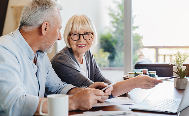 This is a split-screen photograph showing two different scenes. On the left, there s an older couple sitting at a dining table with a laptop and papers, looking attentively at something on the screen. The man appears to be writing, while the woman watches. On the right side of the image, another older couple is shown in a more casual setting, with one person holding a tablet and the other looking at it. They are seated at a table, smiling, and seem to be engaged in a discussion or sharing information on the device. The overall theme suggests a comparison or contrast between work-related activities and leisurely ones involving technology.