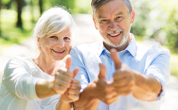 A man and woman giving thumbs-up signs in front of a park with trees.